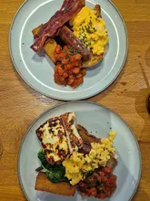 A high-angle, flat-lay photograph of two breakfast plates served on a wooden table. One plate features a traditional full English breakfast with bacon, sausage, scrambled eggs, and beans, while the other is a vegetarian option featuring halloumi, spinach, scrambled eggs, and beans.