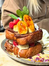 A high-quality, close-up shot of a decadent stack of French toast topped with caramelized bananas, fresh raspberries, and mint, served with a side of whipped cream. The image is well-lit and captures the texture of the food effectively, making it highly appealing for a brunch setting.
