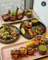 A high-angle, well-composed shot of three distinct Indian dishes served on rectangular and circular plates, featuring grilled lamb chops, a fish/prawn dish, and a samosa chaat, all garnished with fresh herbs and served with dipping sauces on a wooden table.