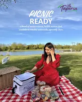 A bright, outdoor lifestyle shot featuring a woman enjoying a picnic by a lake, showcasing a range of Mediterranean-style dips and hummus products from La Taperia.
