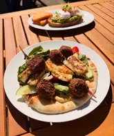 A bright, casual brunch shot featuring a falafel and halloumi flatbread in the foreground and an avocado toast dish in the background, captured on an outdoor wooden table.