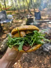 A close-up, handheld shot of a vegan breakfast bagel featuring sausages, bacon, and rocket, taken in an outdoor camping setting.