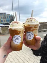 A first-person perspective shot of two hands holding branded Biscoff iced lattes in plastic takeaway cups, positioned against an outdoor background in South Shields.