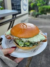 A handheld, close-up shot of a chicken caesar burger held in front of a food truck, highlighting the texture of the breaded chicken and the creamy sauce.