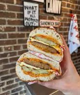 A close-up, hand-held shot of two halves of a breakfast sandwich stacked on top of each other, showcasing the layers of chicken sausage, egg, hash brown, and sauce against a brick wall background.