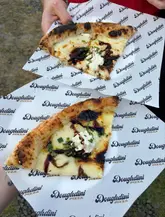 A high-quality, overhead shot of two pizza slices held by a person, served on branded 'Doughdini Pizza' paper. The pizza features a charred crust, melted cheese, and a balsamic glaze drizzle, presenting a fresh and appetizing street food aesthetic.