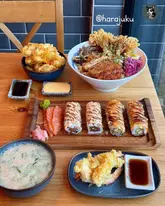 A flat-lay photograph of a Japanese meal spread, featuring sushi rolls, a large rice bowl, tempura shrimp, and a bowl of soup on a wooden table.
