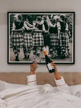 A first-person perspective shot taken from a hotel bed, showing a person holding a bottle of wine and two empty glasses up towards a large framed photograph on the wall.