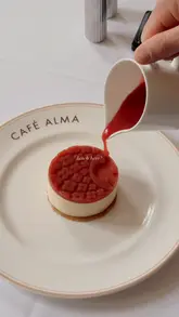 A short, aesthetic montage showcasing the interior and food offerings at Café Alma within the Louis Vuitton Hotel in London. The video features close-ups of a monogrammed dessert, the elegant dining room, and branded coffee.