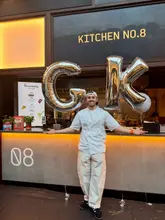 A chef stands in front of his kitchen stall, 'Giuseppe's Kitchen,' holding silver balloons spelling 'GK' to mark the end of a six-week pop-up residency.