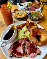 A high-angle shot of a Sunday roast spread at a restaurant, featuring a main plate of roast beef, Yorkshire pudding, roast potatoes, and greens, accompanied by a Bloody Mary and side dishes including cauliflower cheese and aligot mash.