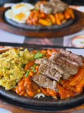 A close-up, high-angle shot of a sizzling steak breakfast platter featuring sliced steak, scrambled eggs, masala beans, and masala chips, with a second plate visible in the blurred background.