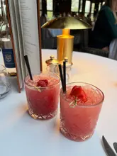 A high-quality, atmospheric shot of two cocktails served on a white-clothed table, likely at a high-end hotel bar. The warm lighting from the table lamp creates an inviting, intimate mood, while the shallow depth of field keeps the focus squarely on the drinks.