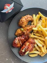 A top-down, casual shot of a plate featuring breaded chicken strips and glazed wings alongside a portion of fries, with a branded takeaway box visible in the corner.