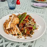 A high-quality, well-lit photograph of a chicken and grain salad served on a white plate, captured outdoors on a patterned terrace table.