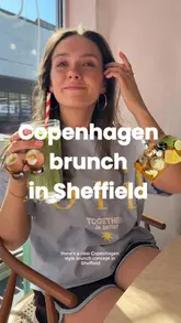 The video features a creator visiting a cafe in Sheffield that offers a 'Copenhagen style' brunch concept. The creator walks the viewer through the ordering process using a tick-sheet menu, showcases the variety of small plates ordered (including eggs, sourdough, avocado, sauerkraut, and pancakes), and highlights the aesthetic interior of the venue.
