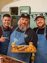 A high-quality, professional portrait of three men wearing branded denim aprons, holding a large, shared tray of BBQ food. The image is well-composed, featuring a clean, on-brand background that suggests a catering or street food environment.