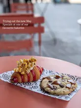 A close-up, eye-level shot of two bakery items—a croissant topped with popcorn and a chocolate chip cookie—resting on branded paper on an outdoor table.