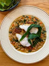 A top-down, well-composed shot of a rustic fish dish served in a white bowl with a gold rim, featuring white fish, greens, and a bean-based stew, set on a wooden table.
