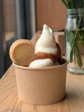 A high-quality, close-up shot of a soft-serve ice cream dessert served in a paper cup, drizzled with caramel sauce and accompanied by a shortbread biscuit. The image is captured with a shallow depth of field, highlighting the texture of the ice cream against a soft, blurred background.