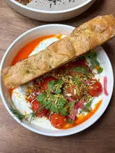 A high-quality, overhead food photography shot of a Turkish-inspired brunch dish featuring herb and garlic yogurt, chilli oil-roasted tomatoes, fresh herbs, and toasted focaccia. The lighting is bright and natural, highlighting the vibrant colors and textures of the meal.