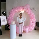 A personal, lifestyle-focused image capturing a birthday celebration held at a Pilates studio. The subject is posed in front of a balloon arch and a cake pedestal, showcasing the venue's event setup capabilities.