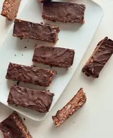 A top-down, well-lit photograph of homemade chocolate oat slices arranged on a white plate. The image captures the texture of the chocolate topping and the oat base, presenting a clean, minimalist aesthetic suitable for food blogging or social media content.