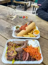 An overhead shot of South African street food, including a boerewors roll and a mixed grill platter, served in plastic takeaway containers on a wooden picnic table with a glass of white wine.