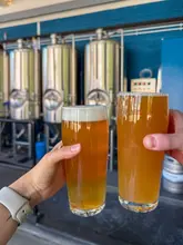 A casual, first-person perspective shot of two glasses of beer being held up in a 'cheers' gesture against the backdrop of brewery fermentation tanks.