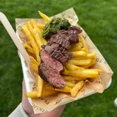 A close-up, top-down shot of a steak and fries meal served in a branded takeaway container, held by a person outdoors at a street food market.