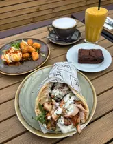 A high-quality, overhead flat-lay shot of a brunch spread at a cafe, featuring a chicken shawarma flatbread, halloumi bites with poached eggs, a coffee, a smoothie, and a brownie, all set on an outdoor wooden table.