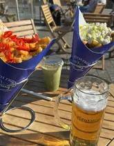A casual, outdoor shot featuring two cones of Dutch fries, a matcha drink, and a lager, captured at a sunny waterside venue.