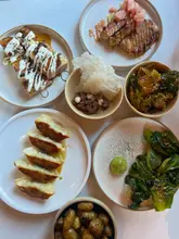 A clean, overhead flat-lay photograph showcasing a variety of Japanese small plates, including gyoza, skewers, cabbage, steak tartare, and a sorbet dessert, arranged on a white table.