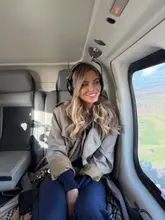 A high-quality, well-composed portrait of a woman inside a helicopter, looking out the window at the landscape below. The lighting is soft and flattering, highlighting the subject clearly against the backdrop of the flight.