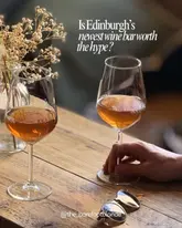 A high-quality, atmospheric shot of two glasses of orange wine on a wooden table, featuring a hand holding one of the glasses. The image uses soft, warm lighting to evoke a cosy, inviting European neighbourhood wine bar feel, effectively capturing the essence of the venue.
