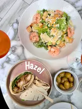 A bright, overhead flat-lay photograph of a lunch spread at Hazel restaurant, featuring a prawn Caesar salad, a southern fried chicken wrap, a bowl of olives, and non-alcoholic spritzes arranged on a marble table.