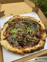 A top-down, eye-level shot of a Hoisin Duck pizza served in a cardboard takeaway box. The pizza features a wood-fired crust, dark sauce, cucumber ribbons, spring onions, and sesame seeds.