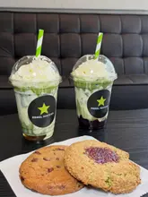 A promotional shot for Rebel Matcha featuring two matcha lattes topped with whipped cream and two cookies served on a piece of paper. The image is taken from a slightly elevated angle against a dark leather sofa background.