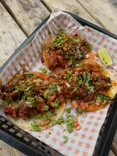 A top-down, overhead shot of three tacos served in a black plastic basket lined with checkered paper, placed on a wooden table. The tacos are topped with fresh cilantro, diced tomatoes, onions, and a chili oil or salsa topping, with a lime wedge on the side.