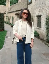 A high-quality portrait of a creator standing in front of a historic stone building, identified as The Wild Duck in the Cotswolds. The image serves as a lifestyle and travel shot, highlighting the venue's exterior architecture and the creator's outfit in a professional, well-composed manner.