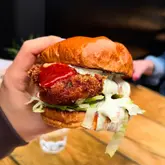 A close-up, handheld shot of a buffalo chicken burger being held up against a blurred background, showcasing the crispy chicken, sauce, and soft bun.