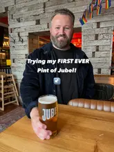 A man poses in a pub holding a pint of Jubel beer, with text overlay announcing his first time trying the drink.