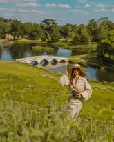 A high-quality, aesthetically pleasing photograph of a woman posing in a large, scenic park with a lake and bridge in the background. The image captures a serene, 'Bridgerton-esque' atmosphere, highlighting the natural beauty of the location.