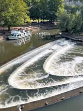 A high-angle, scenic photograph of the Pulteney Weir in Bath, capturing the curved water feature and a tour boat navigating the river. The lighting is bright and natural, highlighting the architecture and the water's movement.