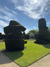 A bright, sunny photograph of the manicured gardens at Billesley Manor, featuring large, sculpted topiary bushes flanking a central white gazebo under a blue sky.