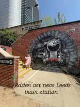 A bright, clear, and well-composed photograph featuring a young girl standing in front of a large, detailed steam train mural near Leeds train station. The natural lighting and clean composition make for an engaging travel-style image.