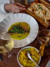 A high-quality, top-down shot of a sophisticated Indian dining experience, featuring a beautifully plated fish dish with a vibrant green sauce, accompanied by rice, curry, and naan, with a glass of white wine in the foreground.