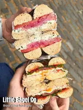 A high-quality, appetizing close-up shot of two stacked, halved bagels held in hands, showcasing the generous fillings and cross-sections against a cobblestone street background.