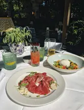 A bright, inviting, and well-composed shot of an outdoor terrace dining setup featuring a burrata salad and a meat dish, accompanied by a cocktail and a drink, capturing the essence of al fresco dining at a hotel venue.