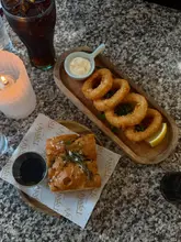 A top-down, atmospheric shot of a restaurant table featuring a wooden serving board with calamari rings and a side of focaccia on branded paper. The scene is lit by a candle, creating a warm, intimate dining environment.
